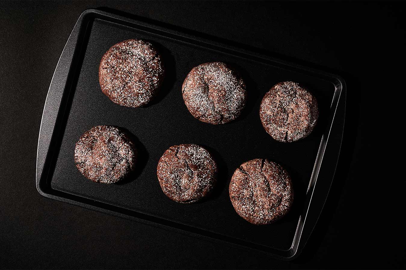 Six chocolate cookies sprinkled with powdered sugar on a black cookie sheet.