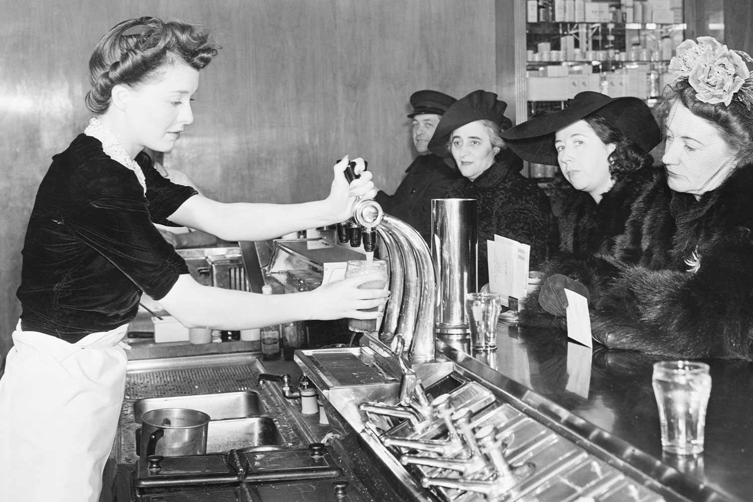 One woman serving milkshakes, and three women and a man seated at a milkshake bar, depicted in black and white. 