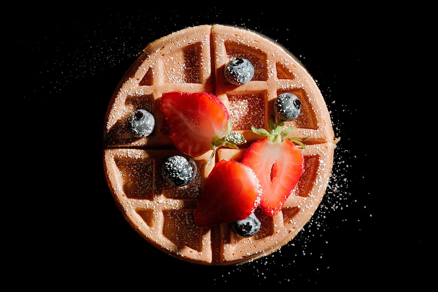 A golden brown waffle topped with strawberries and blueberries on a black background.