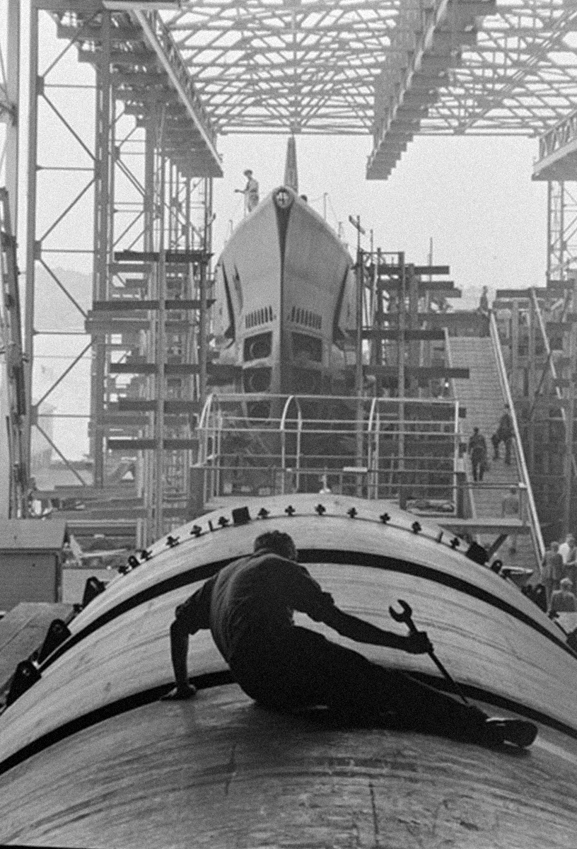 A man sitting on a metal boat part, with scaffolding, a boat bow, and distant figures in the background, depicted in black and white.